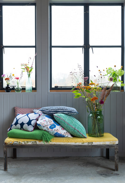 Kitty-Holmes-colourful-and-patterned-cushions-on-yellow-bench-and-vase-of-dried-flowers